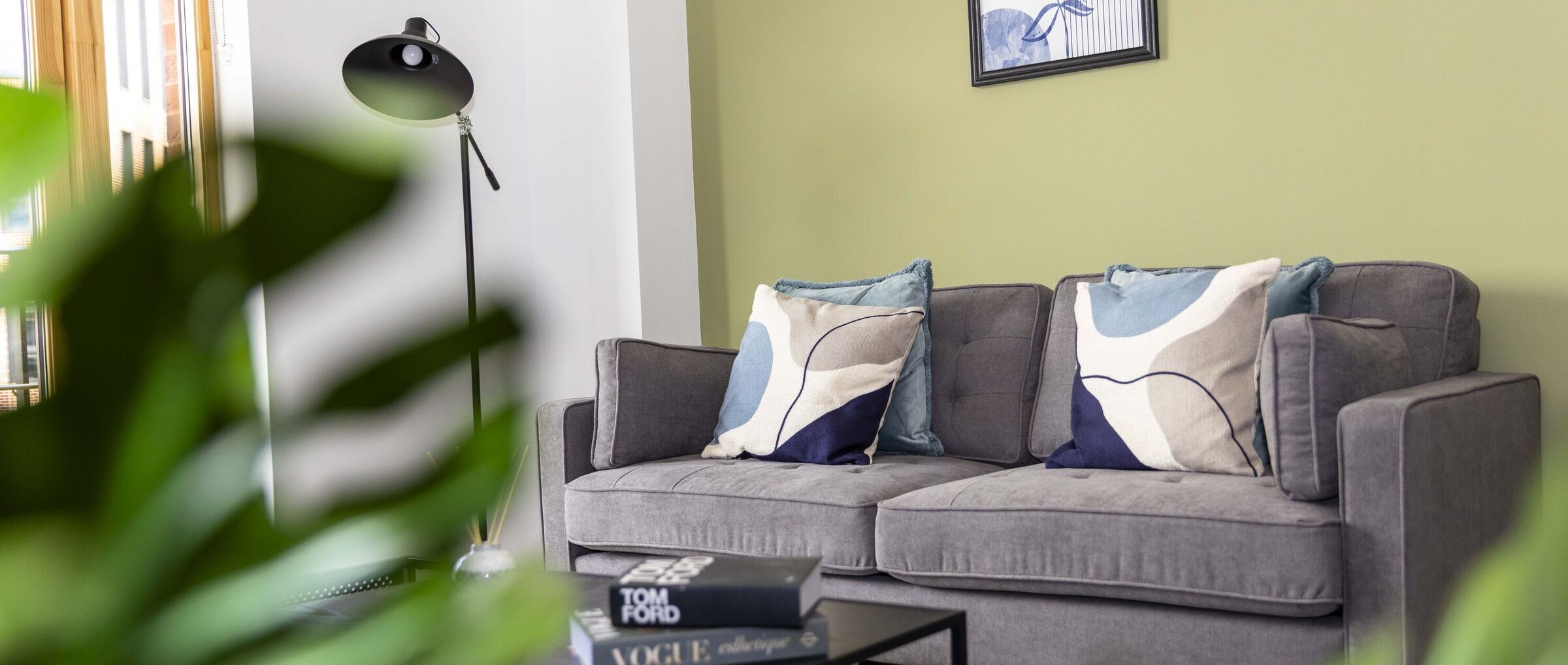 A photograph showing the furnished living room, inclusing a sofa and a coffee tables with some books placed upon it.