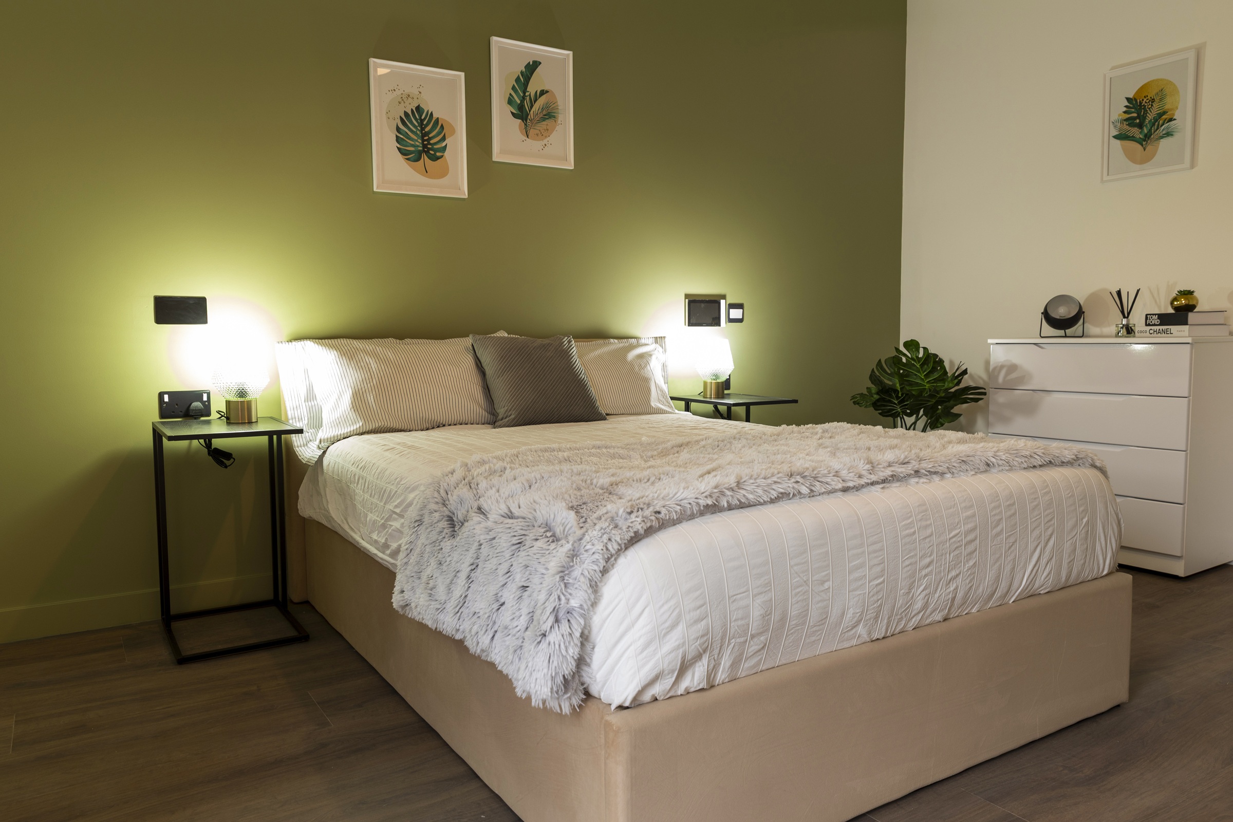 A photograph showing a bedroom furished with a chest of drawers, double bed, side tables, lamps and wall art.